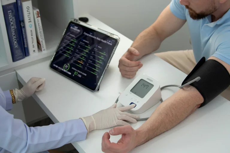 Nurse measuring blood pressure with Omron monitor during TRT Bangkok comprehensive male health check-up — iPad displays full hormonal, metabolic and vitals lab panel with 87/100 overall health score